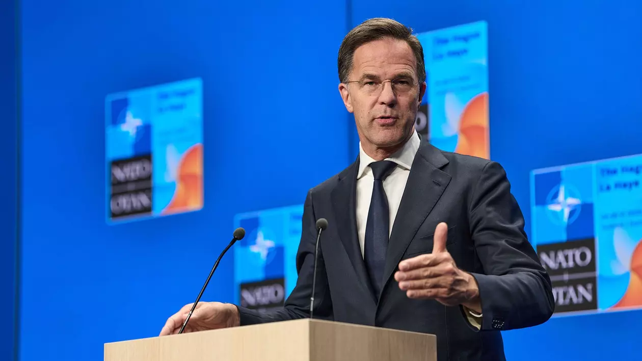Mark Rutte surprins în timpul unei conferințe de presă la summitul NATO. Acesta are o mână ridicată și poartă un sacou negru, o cămașă albă și o cravată neagră