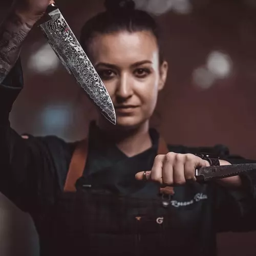 O femeie bucătar, îmbrăcată în negru, ține niște cuțite în mână