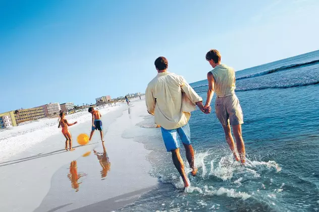 Destinații externe, pentru un city break de 1 Mai . Doi părinți, ținându-se de mână pe malul mării, alături de copiii lor care se joacă cu o minge