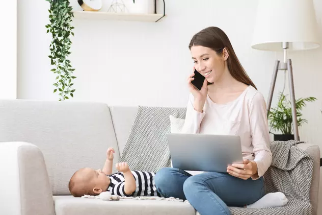 Acte necesare indemnizație creștere copil în 2023- Mama alaturi de nou nascut, stand pe canapea, cu laptopul in fata si telefonul la ureche