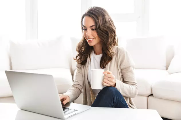 Cursuri online gratuite- Femeie satena cu parul lung, cu cafea in mana, in fata laptopului