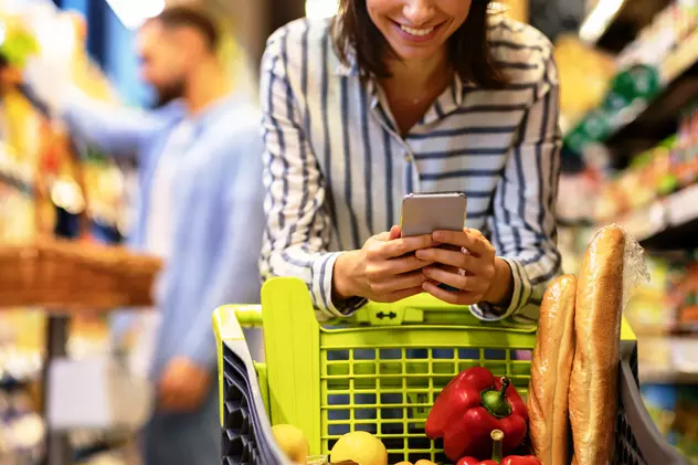 Aplicatii reduceri alimente expirare- O femeie se afla la cumparaturi si sta sprijinita pe cosul de cumparaturi in timp ce se uita in telefon