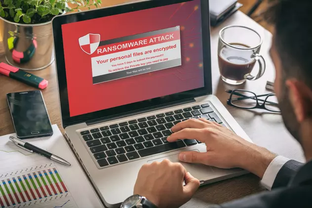 Atacuri Ransomware - Laptop cu imaginea unui atac Ransmoware pe ecran, pus pe un birou, alături de o ceaşcă de cafea, o pereche de ochelari şi obiecte de birotică, agendă, pix, cariocă.