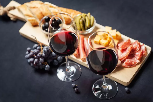 Câte tipuri de vin roșu există - Două pahare de vin roşu alături de un ciorchine de strugure roşu şi un platou cu mezeluri, pe o masă.