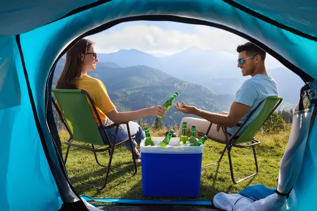 Top 10 cele mai ieftine lăzi frigorifice de voiaj - Un tânăr şi o tânără aflaţi la munte stau pe scaune de voiaj în faţa unui cort, având alături o ladă frigorifică în care se află sicle de bere.