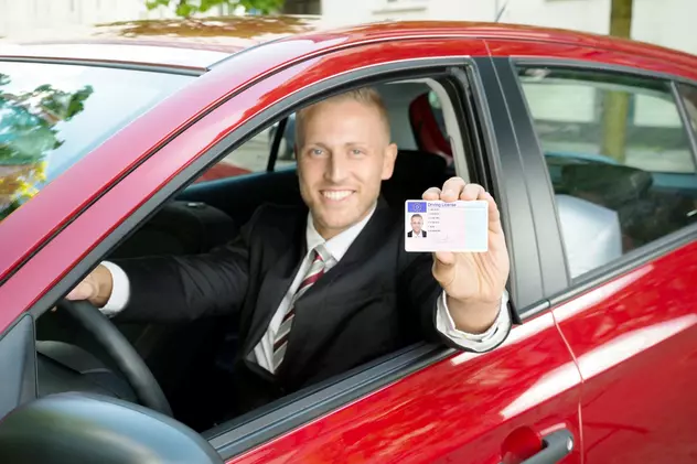 Un barbat aflat la volanul unei masini de culoare rosie isi arata mandru permisul