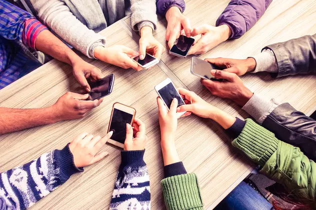 Cât costă iPhone 14 şi Samsung S22 la eMAG înainte de Black Friday - Imagine cu mâinile a şase tineri în jurul unei mese, "butonând" smartphone-uri