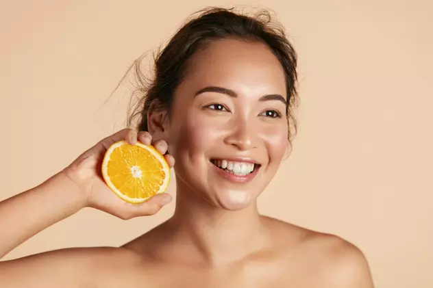 10 motive pentru care vitamina C ar trebui inclusă în rutina ta de îngrijire a pielii - Imagine cu o tânără care ţine lângă faţa ei o jumătate de portocală, sugerând beneficiile vitaminei C în îngrijirea pielii