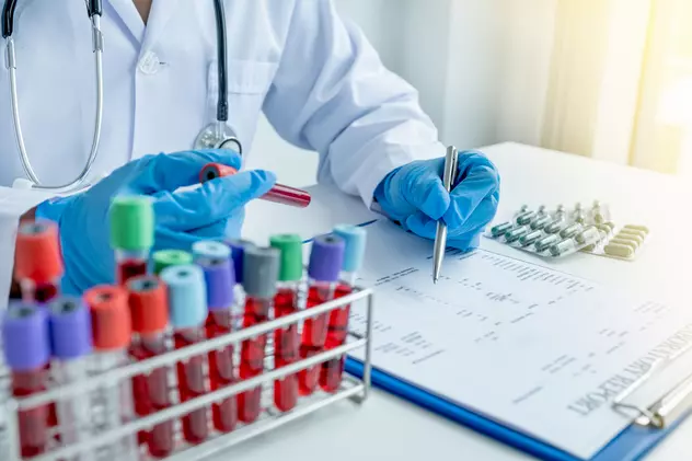 Care este diferența dintre consult și control medical - Imagine cu un medic care notează într-un tabel date de pe flacoanele de sânge pe care le are pe birou, alături de câteva folii de medicamente.