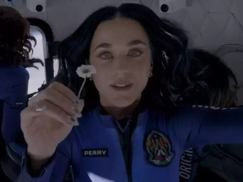 Cântăreața Katy Perry, îmbrăcată în costum albastru, ține o floare mică, albă, în mână, părul îi plutește, în spatele ei alte femei