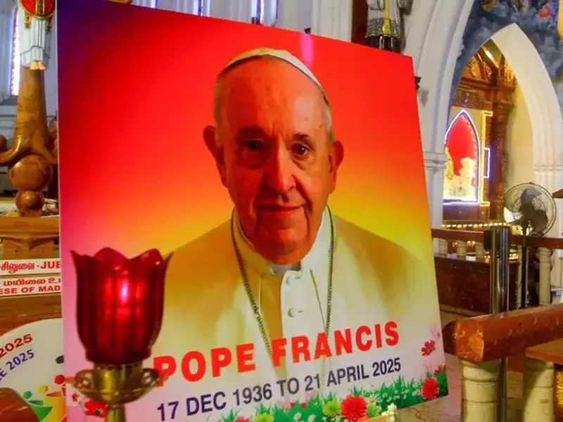 Un portret al Papei Francisc, cu scris pe el și o candelă aprinsă, o statuie a lui Isus Cristos în spate, într-o biserică