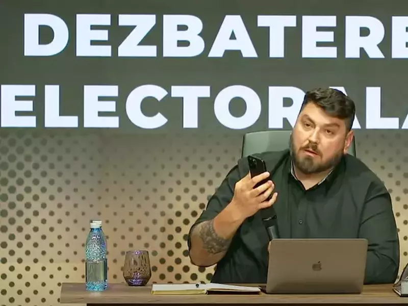 Micutzu alais Cosmin Nedelcu, in studio, la birou, cu un telefon in mana si un laptop in fata