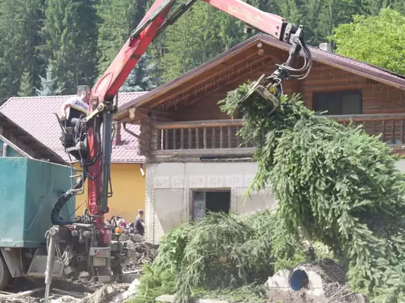 Un exacavator în timp ce ridică brazii pe care furtuna i-a răsturnat și i-a cărat viitura pe străzile din Broșteni, bradul foarte înalt a blocat o curte întreagă a unei case din beton