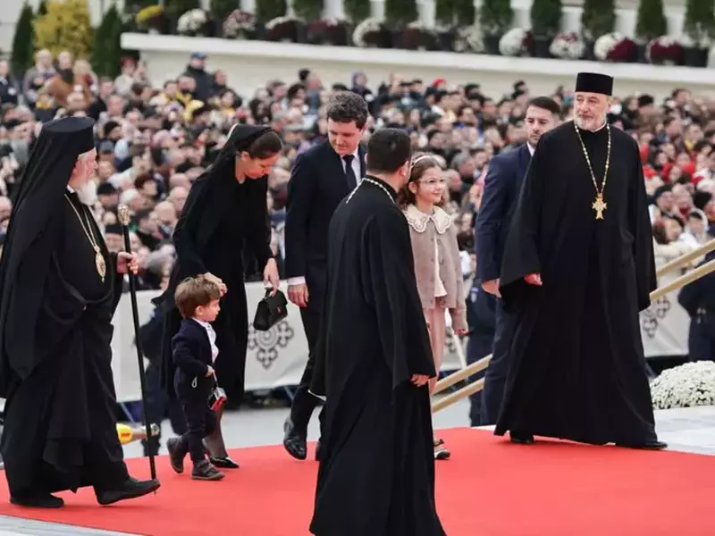 Nicușor Dan, îmbrăcat într-un costum negru, cu cămașă albă cu o cravată neagră, alături de Mirabela Grădinaru, îmbrăcată într-o rochie neagră cu capul acoperit și de copii lor, Patriarhul Daniel și Patriarhul Bartolomeu, înconjurați de soborul de preoți, în timpul ceremoniei de sfințire a picturii Catedralei Mântuirii Neamului, 26 octombrie 2025, București
