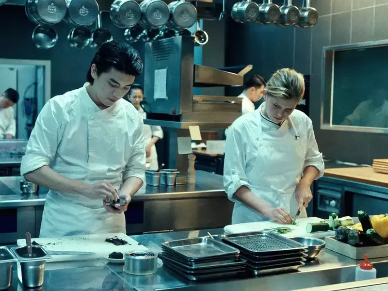 Doi bucatari, un tanar si o tanara, imbracati in alb, gatesc pe o masa intr-un restaurant