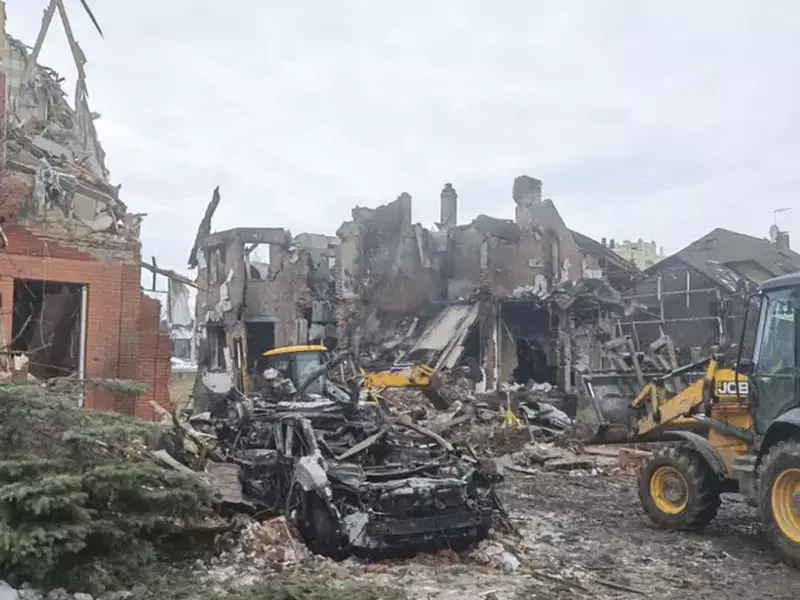 o scenă de distrugeri masive în exterior, unde două clădiri din cărămidă, situate în stânga și în dreapta cadrului, sunt parțial prăbușite, lăsând la vedere structurile interioare de lemn și beton. Spațiul dintre ele este acoperit de un morman mare de moloz compus din resturi de zidărie, grinzi rupte și bucăți de izolație, iar în centrul acestui perimetru se află un buldoexcavator galben parțial vizibil, Borșceahivka, 23 februarie 2026