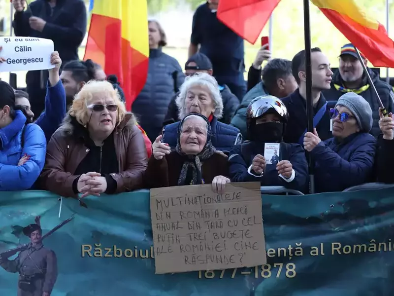 susținătorii lui Călin Georgescu, în fața ICCJ, 28 aprilie 2026, cu steagul României și un carton pe care scrie „Multinaționalele din România pleacă anual din țară cu cel puțin trei bugete ale României. Cine răspunde”
