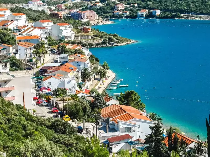 Imaginea prezintă o vedere panoramică a unei localități de coastă, cu numeroase case albe cu acoperișuri roșii dispuse în trepte pe un versant împădurit ce coboară spre apele turcoaz ale unui golf liniștit.