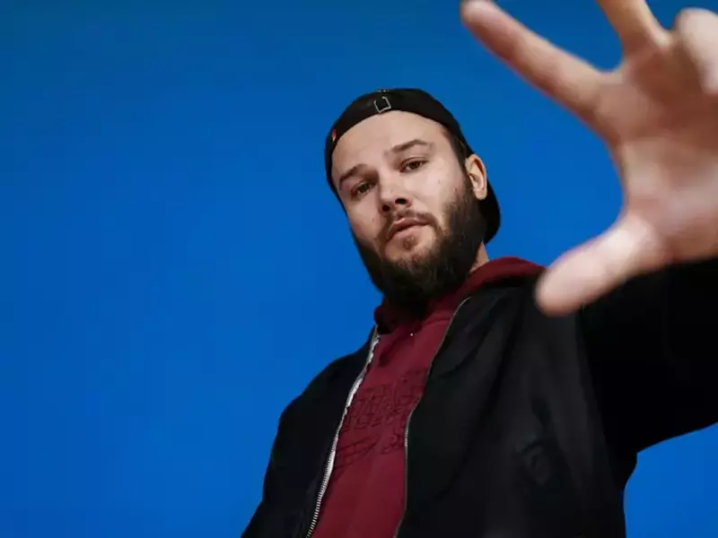 Artistul bielorus Max Korzh, are un hanorac negru și un tricou roșu, în fundal are un perete albastru