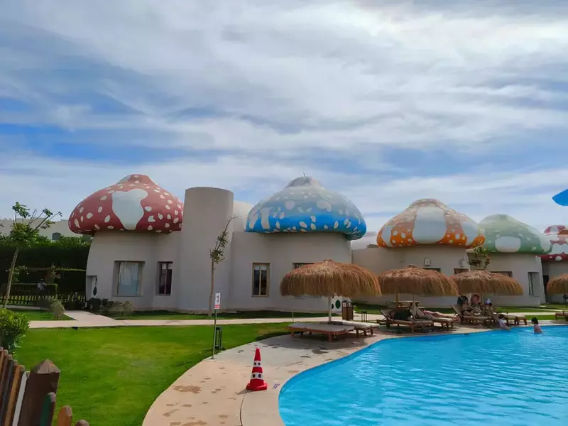 un complex exterior cu o piscină curbată și clădiri cu o arhitectură neobișnuită, având acoperișuri în formă de ciuperci uriașe decorate cu pete, în timp ce pe marginea apei sunt așezate șezlonguri și umbrele cu acoperiș din paie
