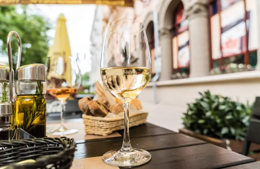 Ce este vinul pelin şi cum se obţine - Imagine cu două pahare de vin alb, un coş cu pâine şi o olivieră, pe o masă de restaurant.