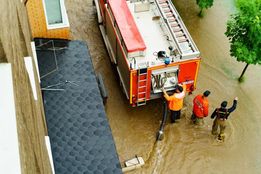 Cum să acţionezi în caz de inundaţii - Imagine cu pompieri şi o maşină de pompieri care intervin pentru îndepărtarea efectelor unei inundaţii într-o zonă urbană.