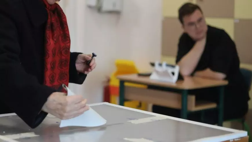 Femeie care poartă o bluză neagră și o eșarfă roșie cu model cu flori care introduce un buletin de vot îndoit în urnă
