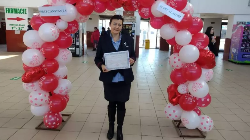Femeie în fața unei arcade de baloane albe și roșii cu un certificat în mână în Gara Constanța