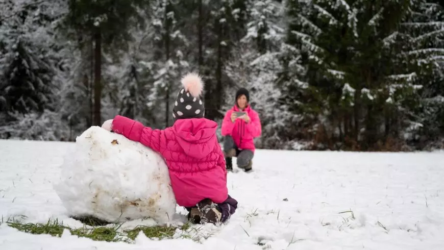 Un copil mic într-o haină roz de iarnă rostogolește un bulgăre mare de zăpadă, în timp ce un adult din fundal face o fotografie, surprinzând un moment de bucurie într-o pădure înzăpezită Sursă Profimedia