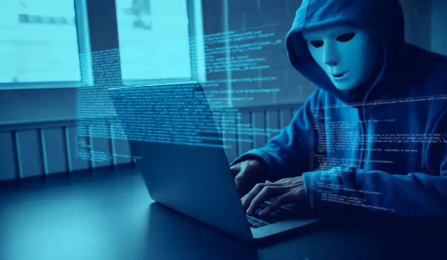 bărbat cu glugă și cu o mască albă pe față, care stă pe laptop. imaginea este acoperită de un text tip cod informatic și este colorată în lumină albastră