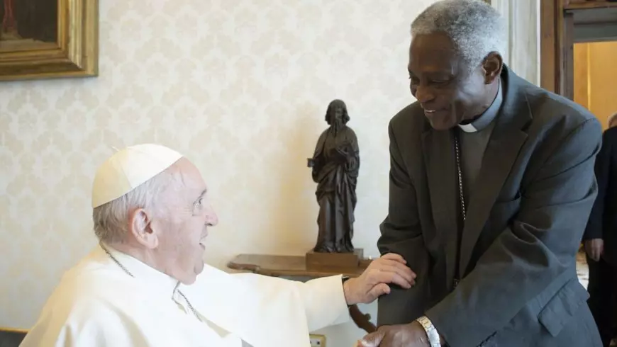 Papa Francis, asezat, înbrăcat în alb, il tine de mâna pe Cardinalul Turkson, în picioare, îmbrăcat în negru