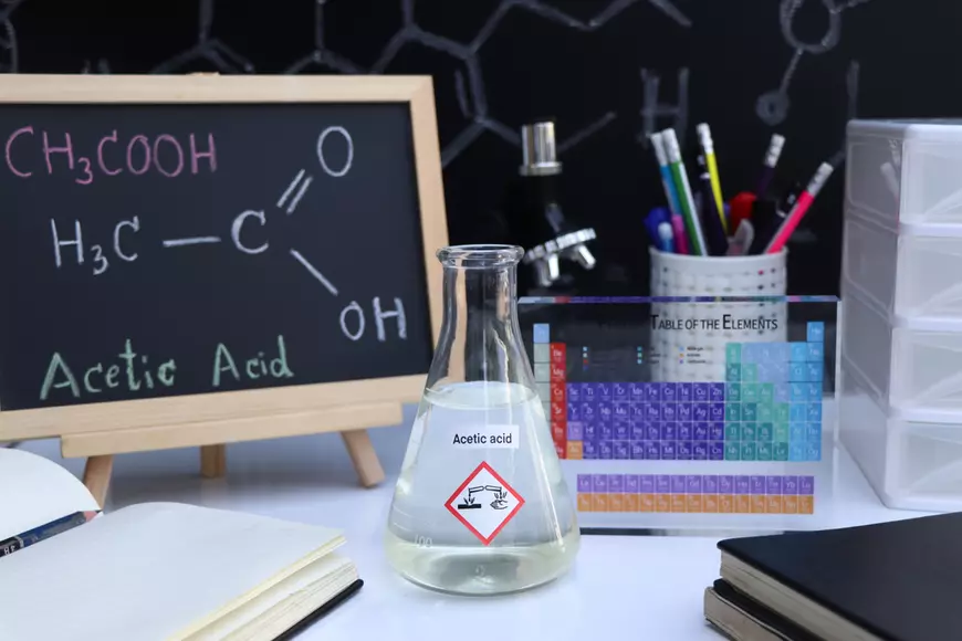 Care este diferenţa dintre oţet şi acid acetic alimentar - Imagine cu un pahar erlenmayer, în care se află acid acetic, aşezat pe un birou, în care se mai află, printre altele, o tâbliţă pe care este scrisă cu creta formua acidului acetic.