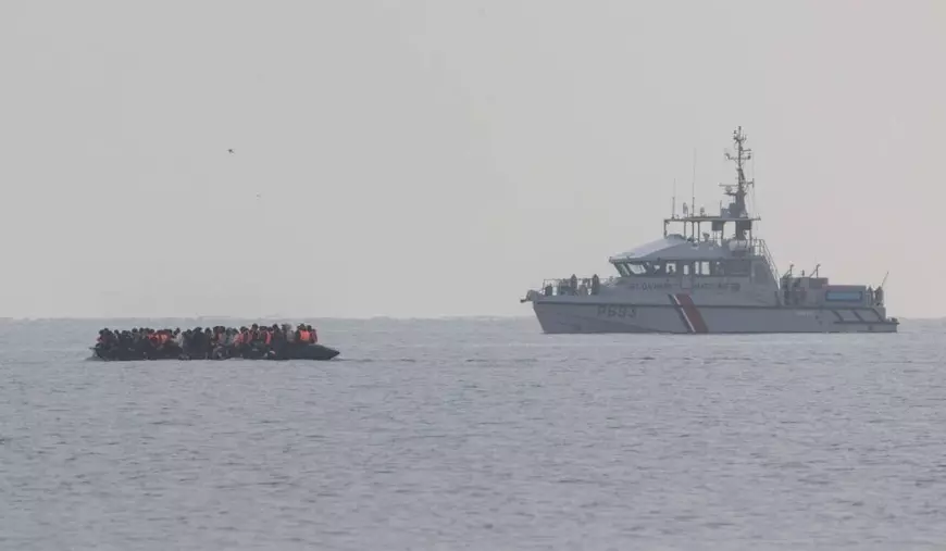 Imigranții ilegali stând pe o barcă gonflabila și fiind transportați pe apă către un vapor. Fiecare cetățean are vestă de salvare.