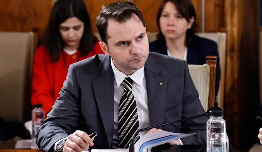 Ministrul Energiei, Sebastian Burduja, în timpul unei ședințe de Guvern, poartă o cravată cu dungi și stă la masă cu agenda deschisă.