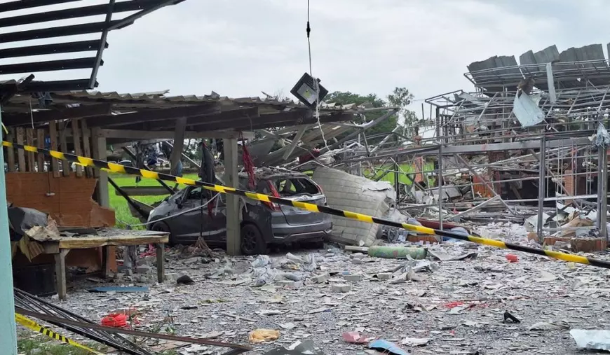 rămășițele unei fabrici de artificii din Thailanda după o explozie - în imaginea se vede o clădire cu acoperiș de tabla de culoare portocalie, înconjurată de funingine și bucăți de lemn și de metal. În fața clădirii este întinsă o bandă de protecție a poliției, cu dungi galbene și negre