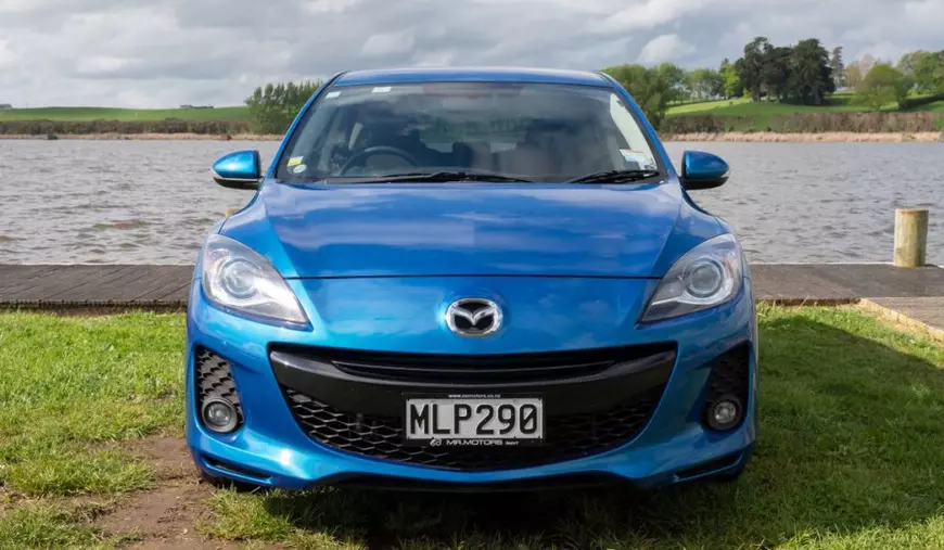 Mazda model 3, de culoare albastru, pe malul unui lac, parcat pe iarbă, fotografiat din față