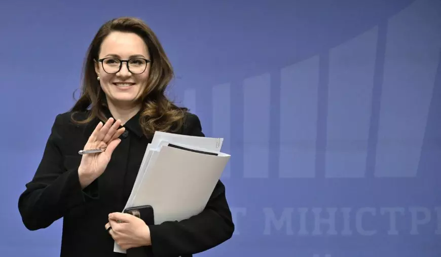 Yulia Svyrydenko, zâmbind larg, îmbrăcată într-un costum negru format din pantaloni și sacou, și cu o bluză neagră, ținând la piept cu mâna stângă niște dosare albe și un telefon cu husă neagră, și ținând mâna dreaptă ridicată, cu palma deschisă, făcând cu mâna fotografilor