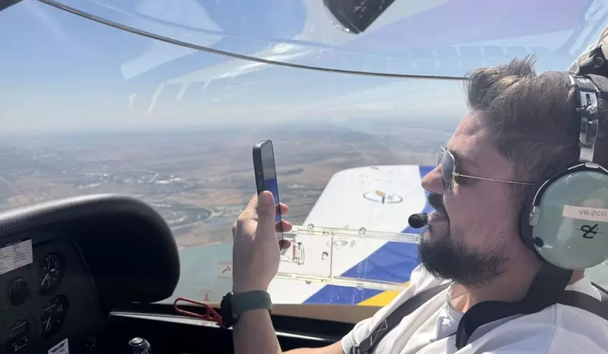 Bărbat care zboară un avion de acrobație, în cockpit, cu căștile pe urechi, telefonul în mână, îmbrăcat într-un tricou alb cu cerul și linia orizontului pe fundal.