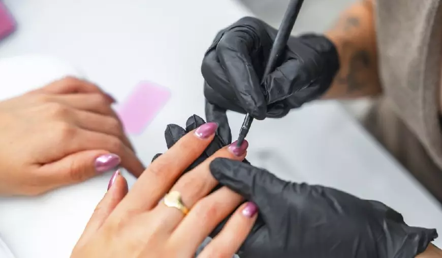 Cosmeticiană purtând mănuși negre aplicând cu grijă lac de unghii pe unghiile clientei, realizând o manichiură perfectă într-un salon de înfrumusețare profesional.