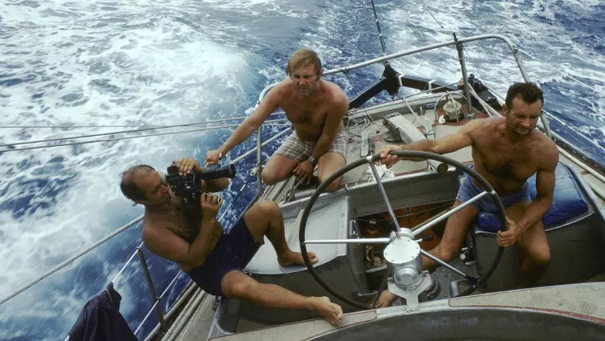 In imagine Eric Tabarly (dreapta) la cârma lui Pen Duick VI, in stanga jurnalistul Christian Brincourt., cu camera in mana, în spate un alt bărbat, toti la bustul gol pe o mare agitată,1978