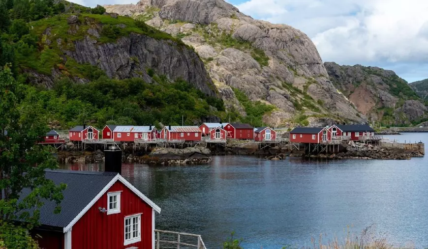 Un grup de cabane roșii vibrante stau pe picioroange de-a lungul țărmului stâncos și liniștit. Satul pescăresc Nusfjord Insulele Lofoten Norvegia