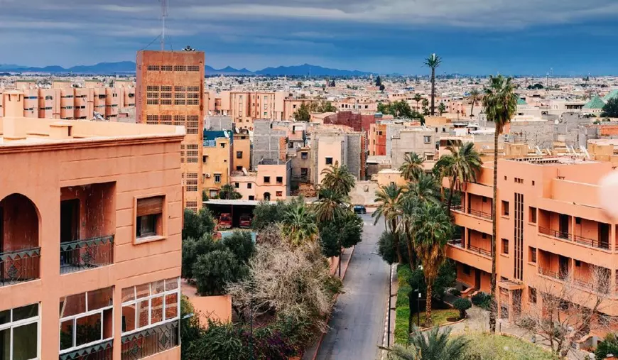 Clădirile în culoarea ocru și palmierii care umplu peisajul urban din Marrakech sub un cer înnorat creează o scenă urbană vibrantă în Maroc.
