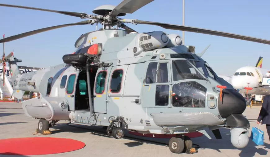 Un elicopter Airbus H225M, aflat la sol, cu palele întinse, pe un aeroport pe care se văd pe fundal avioane comerciale de culoare albă