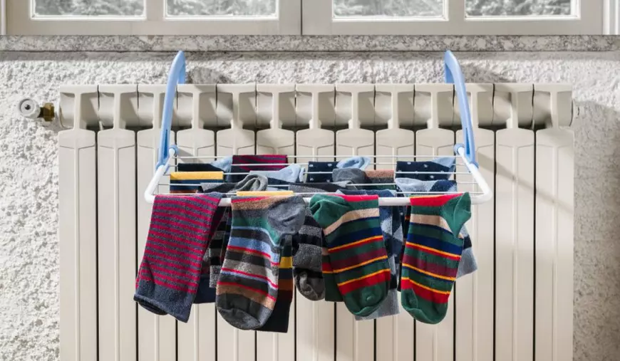 Haine de diferite culori, puse la uscat pe un uscător metalic plasat în fața unui calorifer care se află sub o fereastră