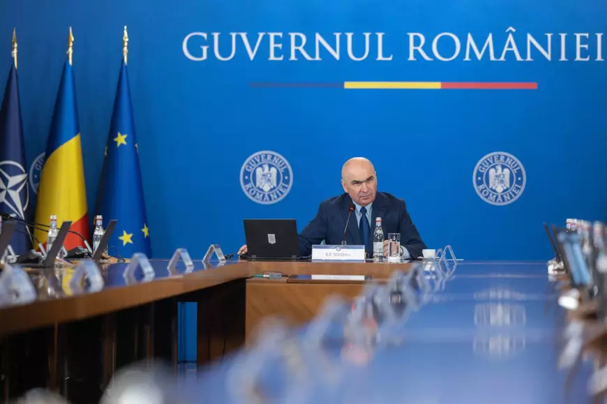 Ilie Bolojan conduce ședința de guvern din 14 ianuarie 2026. În spate se vede scris pe un perete Guvernul României, iar în dreapta lui are 3 steaguri: al României, NATO și Uniunii Europene