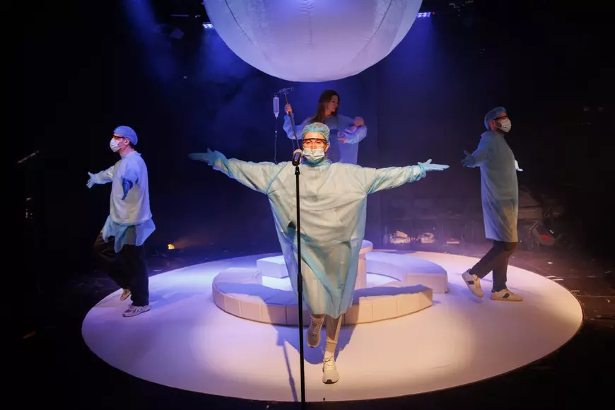 Imagine din spectacolul ”The woman” în care doctorii din spectacol țin mâinile deschise iar pe fundal se vede actrița principală cu un bebeluș în brațe.