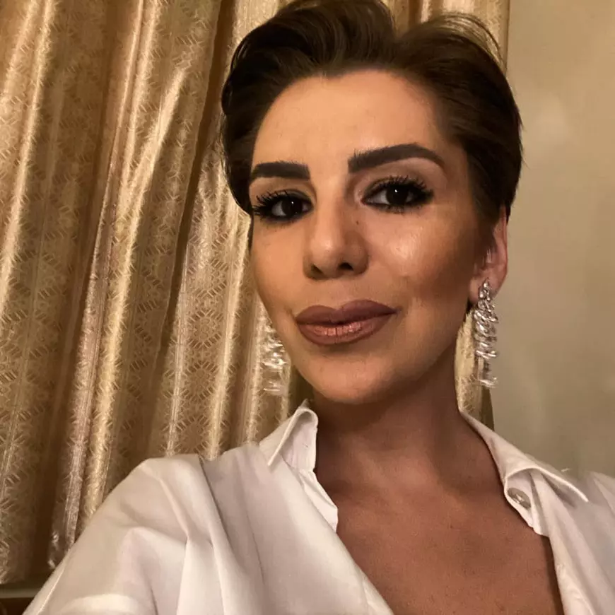 Adriana Viliginschi, îmbrăcată cu o bluză albă, cerceri argintii lungi la urechi, părul scurt, iar pe fundal o perdea aurie