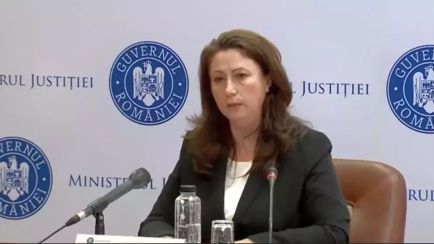 O doamna imbracată în costum negru stă la o masă și vorbește la microfon