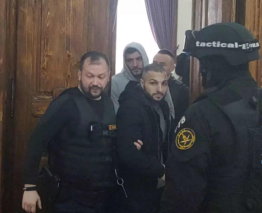 Laurențiu Ghiță și Marian Cristian Minae (în spate, în gri) au fost condamnați pentru uciderea lui Adrian Kreiner. In foto, doi ofiteri de politie duc la audieri un barbat imbracat in negru si unul in gri in spatele celui in negru