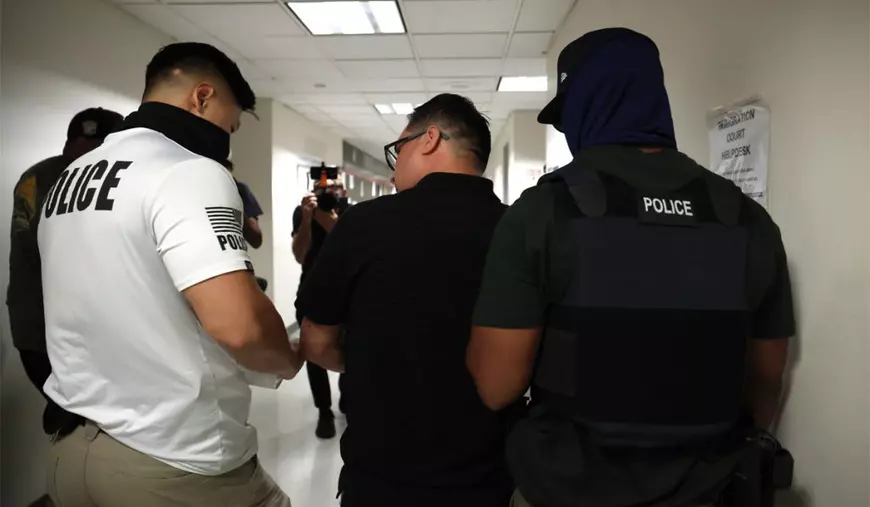 Doi polițiști cu fețele acoperite escortează o persoană arestată pe care vor să o extrădeze.